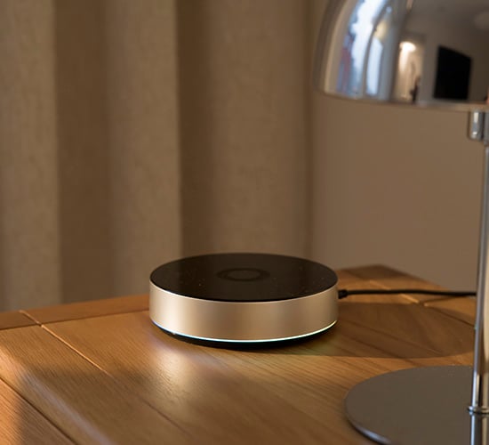 Rund smart hem-hub med svart topp och metallram, placerad på ett träbord med svagt lysande LED-ring i botten.